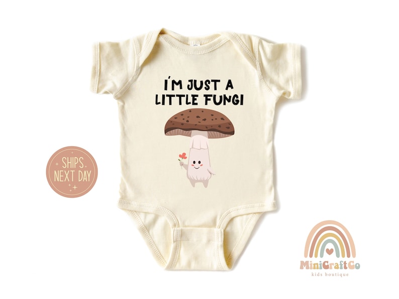 May include: Cream-colored baby bodysuit with the text "I'M JUST A LITTLE FUNGI" above a cartoon mushroom holding a flower. The mushroom has a brown cap and a smiling face. A rainbow logo is in the bottom right.