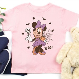 K&ouml;nnte beinhalten: Ein hellrosa T-Shirt mit kurzen &Auml;rmeln und einer Grafik von Minnie Maus in einem lila Kleid mit Schleife, die einen Totenkopf-Lutscher und einen K&uuml;rbis h&auml;lt. Das Design zeigt zudem ein Spinnennetz, Flederm&auml;use, Sterne und das Wort BOO in Dunkelbraun.