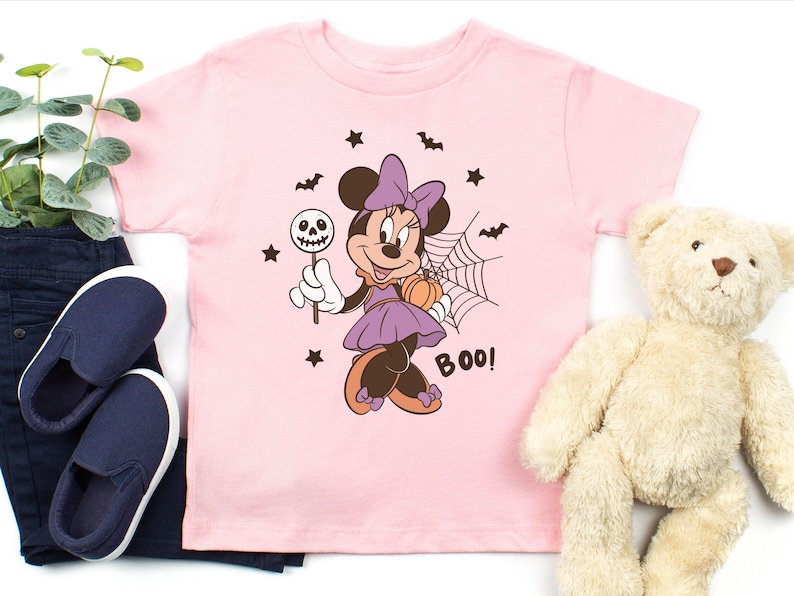K&ouml;nnte beinhalten: Ein hellrosa T-Shirt mit kurzen &Auml;rmeln und einer Grafik von Minnie Maus in einem lila Kleid mit Schleife, die einen Totenkopf-Lutscher und einen K&uuml;rbis h&auml;lt. Das Design zeigt zudem ein Spinnennetz, Flederm&auml;use, Sterne und das Wort BOO in Dunkelbraun.