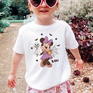 K&ouml;nnte beinhalten: Ein wei&szlig;es Kurzarm-T-Shirt mit einer Grafik von Minnie Maus in einem lila Kleid mit Schleife, die einen Totenkopf-Lutscher und einen K&uuml;rbis h&auml;lt. Das Design zeigt zudem ein Spinnennetz, kleine Flederm&auml;use, Sterne und den Schriftzug BOO!.