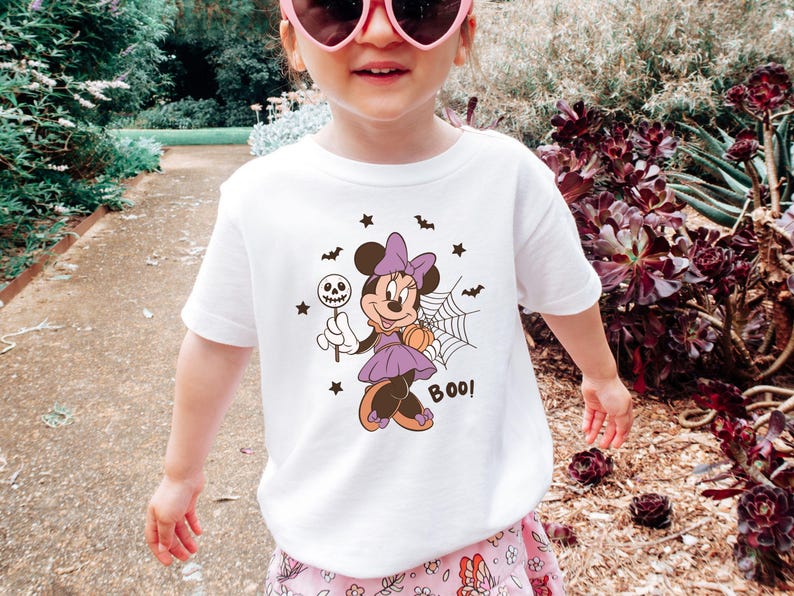 K&ouml;nnte beinhalten: Ein wei&szlig;es Kurzarm-T-Shirt mit einer Grafik von Minnie Maus in einem lila Kleid mit Schleife, die einen Totenkopf-Lutscher und einen K&uuml;rbis h&auml;lt. Das Design zeigt zudem ein Spinnennetz, kleine Flederm&auml;use, Sterne und den Schriftzug BOO!.