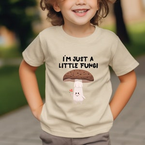 May include: Beige t-shirt with the text "I'M JUST A LITTLE FUNGI" above a cartoon mushroom illustration. The mushroom has a brown cap, white stem, and a smiling face. A small red heart is next to the mushroom.