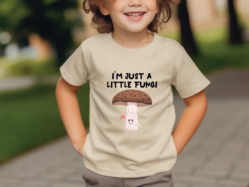 May include: Beige t-shirt with the text "I'M JUST A LITTLE FUNGI" above a cartoon mushroom illustration. The mushroom has a brown cap, white stem, and a smiling face. A small red heart is next to the mushroom.