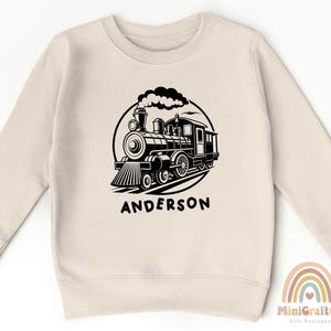 May include: A cream colored sweatshirt with a black and white illustration of a train engine with the name "Anderson" printed below.