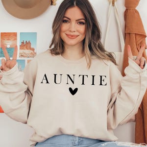 May include: A beige sweatshirt with the word "AUNTIE" printed in black letters and a small black heart below the word.
