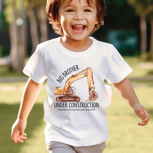 Pode incluir: Camiseta branca com uma ilustra&ccedil;&atilde;o em aquarela de uma escavadeira. O texto diz "IRM&Atilde;O MAIS VELHO EM CONSTRU&Ccedil;&Atilde;O" e "Data estimada de conclus&atilde;o junho de 2024". Usada por uma crian&ccedil;a sorrindo.