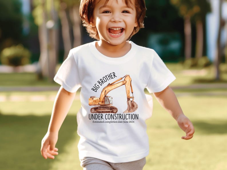 Pode incluir: Camiseta branca com uma ilustra&ccedil;&atilde;o em aquarela de uma escavadeira. O texto diz "IRM&Atilde;O MAIS VELHO EM CONSTRU&Ccedil;&Atilde;O" e "Data estimada de conclus&atilde;o junho de 2024". Usada por uma crian&ccedil;a sorrindo.