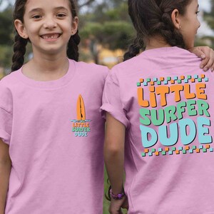 K&ouml;nnte beinhalten: Rosa T-Shirt mit einem Surfboard-Motiv und dem Text "Little Surfer Dude" auf der Vorderseite und dem Text "Little Surfer Dude" in bunten Buchstaben auf der R&uuml;ckseite.