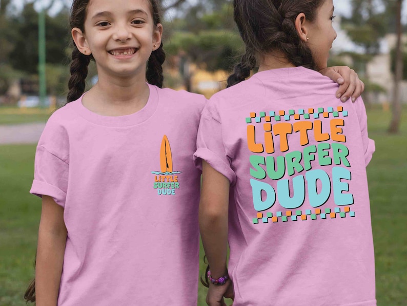 K&ouml;nnte beinhalten: Rosa T-Shirt mit einem Surfboard-Motiv und dem Text "Little Surfer Dude" auf der Vorderseite und dem Text "Little Surfer Dude" in bunten Buchstaben auf der R&uuml;ckseite.