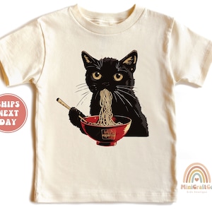 May include: A black cat wearing a white t-shirt with a graphic of a black cat eating ramen noodles from a red bowl with chopsticks.
