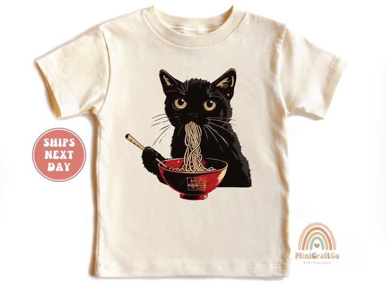May include: A black cat wearing a white t-shirt with a graphic of a black cat eating ramen noodles from a red bowl with chopsticks.