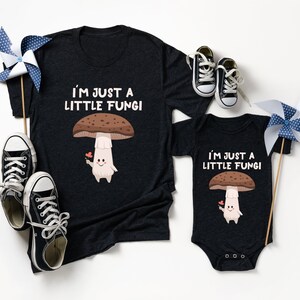 May include: Dark gray t-shirt and onesie set with a mushroom graphic and the text "I'M JUST A LITTLE FUNGI". The mushroom has a brown cap, a white stem, and is holding a flower. Black and white sneakers and pinwheels are also in the image.