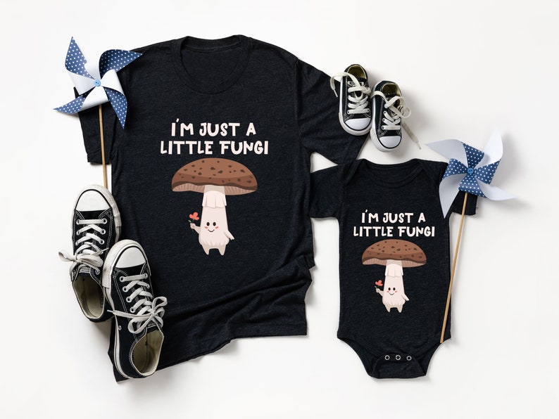 May include: Dark gray t-shirt and onesie set with a mushroom graphic and the text "I'M JUST A LITTLE FUNGI". The mushroom has a brown cap, a white stem, and is holding a flower. Black and white sneakers and pinwheels are also in the image.