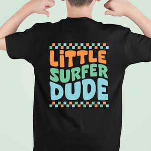 K&ouml;nnte beinhalten: Schwarzes T-Shirt mit dem Text "Little Surfer Dude" in bunten, stilisierten Buchstaben. Der Text ist von einer Reihe kleiner orangefarbener, gr&uuml;ner und blauer Quadrate eingerahmt.