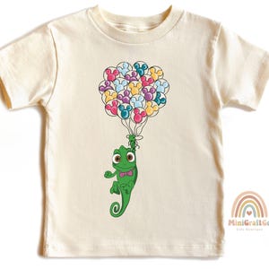 May include: A light beige t-shirt with a green chameleon holding a bunch of colorful balloons shaped like Mickey Mouse ears. The balloons are pink, blue, yellow, green, and purple.