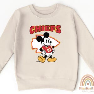 May include: Cream-colored sweatshirt featuring a graphic of Mickey Mouse in a Chiefs football jersey. The word "CHIEFS" is printed above the character in red and yellow. The number "87" is visible on the jersey. The sweatshirt has long sleeves and a crew neck.