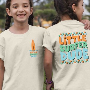 K&ouml;nnte beinhalten: Zwei Kinder tragen wei&szlig;e T-Shirts mit einem Surfer-Motiv. Die Vorderseite des Shirts zeigt ein kleines orangefarbenes Surfbrett mit dem Text "Little Surfer Dude". Die R&uuml;ckseite des Shirts zeigt den Text "Little Surfer Dude" in gro&szlig;en, bunten Buchstaben.