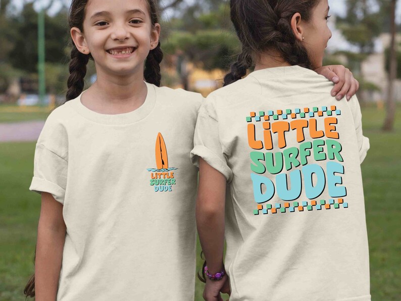 K&ouml;nnte beinhalten: Zwei Kinder tragen wei&szlig;e T-Shirts mit einem Surfer-Motiv. Die Vorderseite des Shirts zeigt ein kleines orangefarbenes Surfbrett mit dem Text "Little Surfer Dude". Die R&uuml;ckseite des Shirts zeigt den Text "Little Surfer Dude" in gro&szlig;en, bunten Buchstaben.