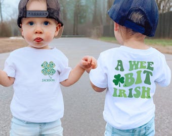 Puede incluir: Dos camisetas blancas con diseños del Día de San Patricio. Una camiseta presenta un trébol verde y amarillo con el nombre "Jackson" debajo. La otra camiseta dice "A Wee Bit Irish" con un trébol verde.