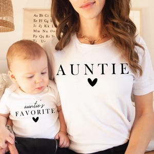 May include: A white t-shirt with the word "AUNTIE" printed in black with a small black heart below. A child wearing a white t-shirt with the text "auntie's FAVORITE" printed in black with a small black heart below.