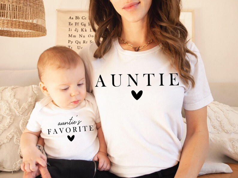 May include: A white t-shirt with the word "AUNTIE" printed in black with a small black heart below. A child wearing a white t-shirt with the text "auntie's FAVORITE" printed in black with a small black heart below.
