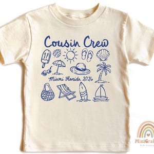 May include: A cream-colored short-sleeve t-shirt featuring a blue graphic print with the text "Cousin Crew" and "Miami Florida 2026". The design includes illustrations of a popsicle, sun, flip-flops, seashell, umbrella, hat, palm tree, bag, chair, surfboard, and sailboat.
