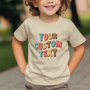 May include: A child wearing a beige t-shirt with the text "YOUR CUSTOM TEXT" in colorful, block letters. The letters are in shades of pink, blue, yellow, and brown.