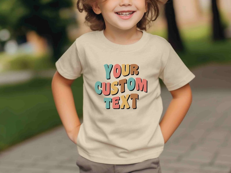 May include: A child wearing a beige t-shirt with the text "YOUR CUSTOM TEXT" in colorful, block letters. The letters are in shades of pink, blue, yellow, and brown.