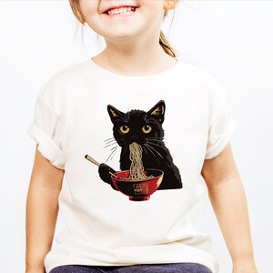 May include: A white t-shirt with a black cat eating noodles from a red bowl. The cat has a serious expression on its face.