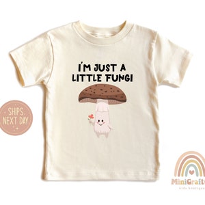 May include: Cream-colored toddler t-shirt with the text "I'M JUST A LITTLE FUNGI" above a cartoon mushroom character holding a flower. The mushroom has a brown cap and a smiling face.