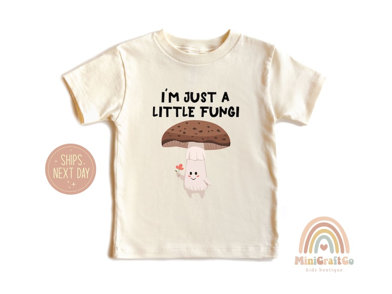 May include: Cream-colored toddler t-shirt with the text "I'M JUST A LITTLE FUNGI" above a cartoon mushroom character holding a flower. The mushroom has a brown cap and a smiling face.