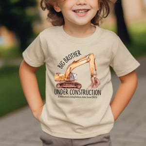 Pode incluir: Camiseta bege com uma ilustra&ccedil;&atilde;o em aquarela de uma escavadeira. O texto diz "Big Brother Under Construction" com uma data estimada de conclus&atilde;o em junho de 2024. Usada por uma crian&ccedil;a.
