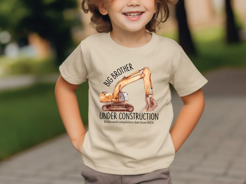 Pode incluir: Camiseta bege com uma ilustra&ccedil;&atilde;o em aquarela de uma escavadeira. O texto diz "Big Brother Under Construction" com uma data estimada de conclus&atilde;o em junho de 2024. Usada por uma crian&ccedil;a.