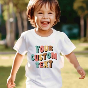 May include: A white t-shirt with a colorful graphic that says "YOUR CUSTOM TEXT". The text is in a retro style font with each letter in a different color. A young child is wearing the shirt and smiling.