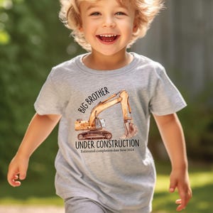 Pode incluir: Uma camiseta cinza claro com uma ilustra&ccedil;&atilde;o em aquarela de uma escavadeira de constru&ccedil;&atilde;o. O texto na camiseta diz "BIG BROTHER UNDER CONSTRUCTION" com uma data de conclus&atilde;o estimada em junho de 2024.