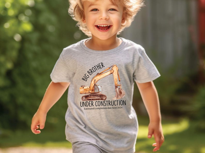 Pode incluir: Uma camiseta cinza claro com uma ilustra&ccedil;&atilde;o em aquarela de uma escavadeira de constru&ccedil;&atilde;o. O texto na camiseta diz "BIG BROTHER UNDER CONSTRUCTION" com uma data de conclus&atilde;o estimada em junho de 2024.
