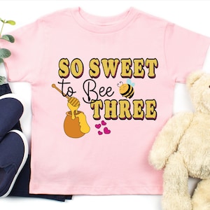 May include: Pink t-shirt with a yellow and black bee graphic and the text "So Sweet to Bee Three".