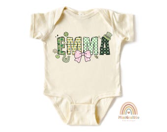 Puede incluir: Un body de bebé de color crema con mangas cortas presenta un diseño estampado en verde, dorado y blanco que deletrea el nombre "EMMA". Un lazo rosa está por encima del nombre y un trébol verde a la izquierda.
