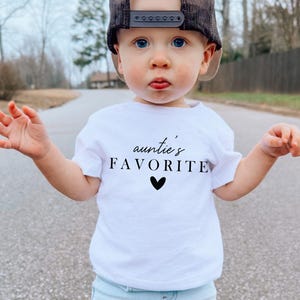 May include: A white t-shirt with black text that says "auntie's favourite" with a small black heart below the text. A young child is wearing the shirt and a black baseball cap.