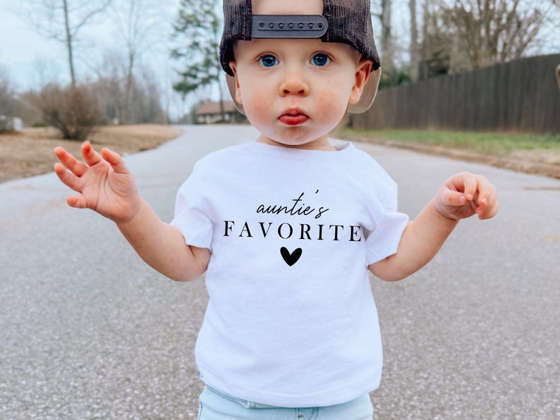 May include: A white t-shirt with black text that says "auntie's favourite" with a small black heart below the text. A young child is wearing the shirt and a black baseball cap.