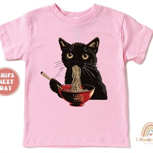 May include: Pink short-sleeved t-shirt with a black cat eating ramen noodles from a red bowl. The cat has yellow eyes and a black nose.