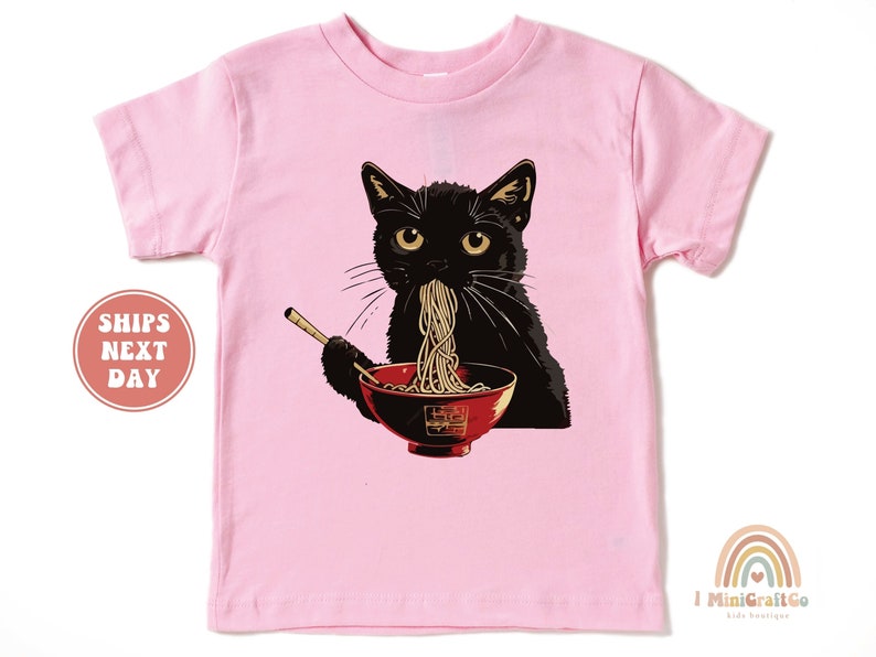 May include: Pink short-sleeved t-shirt with a black cat eating ramen noodles from a red bowl. The cat has yellow eyes and a black nose.
