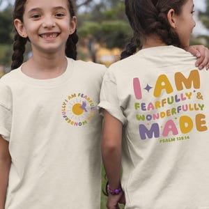 May include: Two children wearing white t-shirts with a colorful graphic design. The front of the shirt says "I am fearfully and wonderfully made" with a sun and bird design. The back of the shirt says "I am fearfully and wonderfully made" with a star and the text "Psalm 139:14".