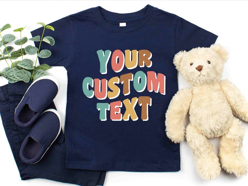 May include: A navy blue t-shirt with the text "YOUR CUSTOM TEXT" in colorful, outlined letters. The letters are in shades of teal, orange, pink, and brown.