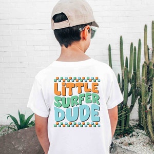K&ouml;nnte beinhalten: Wei&szlig;es T-Shirt mit einer bunten Grafik, auf der hinten "Little Surfer Dude" steht. Das Shirt wird von einer Person getragen, die eine beige Baseballkappe und eine Sonnenbrille tr&auml;gt.