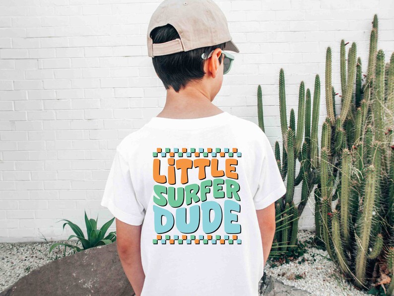 K&ouml;nnte beinhalten: Wei&szlig;es T-Shirt mit einer bunten Grafik, auf der hinten "Little Surfer Dude" steht. Das Shirt wird von einer Person getragen, die eine beige Baseballkappe und eine Sonnenbrille tr&auml;gt.