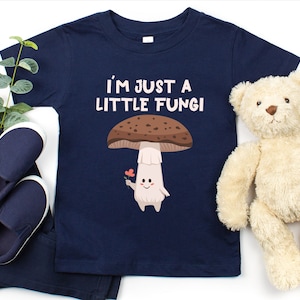 May include: Navy blue toddler t-shirt with the text "I'M JUST A LITTLE FUNGI" and a cartoon mushroom graphic. The mushroom has a brown cap, white stem, and is holding a flower. The shirt is displayed with navy pants, shoes, and a teddy bear.