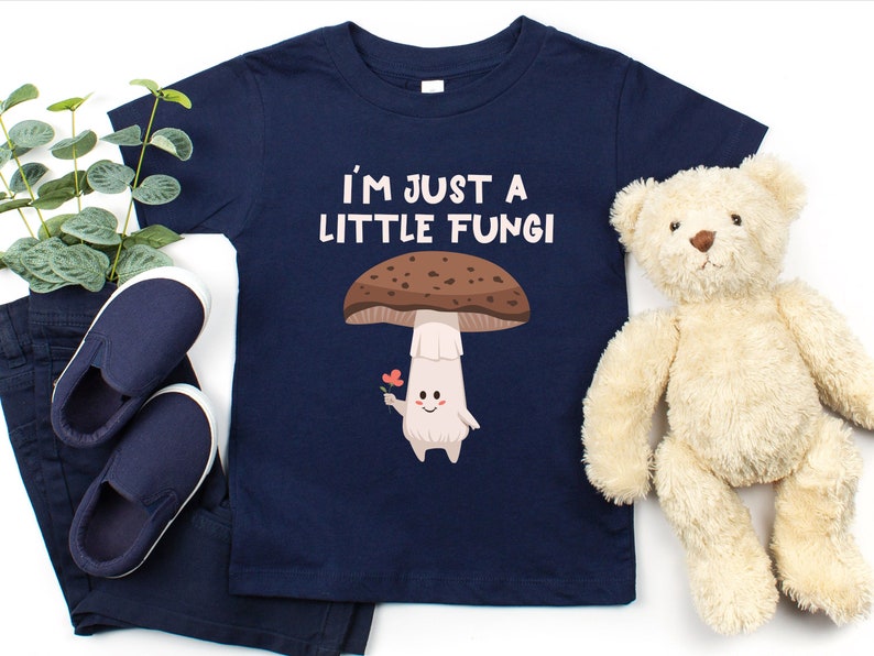 May include: Navy blue toddler t-shirt with the text "I'M JUST A LITTLE FUNGI" and a cartoon mushroom graphic. The mushroom has a brown cap, white stem, and is holding a flower. The shirt is displayed with navy pants, shoes, and a teddy bear.