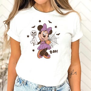 K&ouml;nnte beinhalten: Ein wei&szlig;es T-Shirt mit einer Grafik von Minnie Maus in einem lila Kleid mit passender Schleife, die einen Totenkopf-Lutscher und einen K&uuml;rbis h&auml;lt. Das Motiv zeigt zudem ein Spinnennetz, kleine schwarze Flederm&auml;use, Sterne und das Wort BOO.