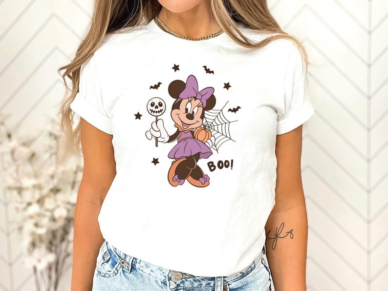 K&ouml;nnte beinhalten: Ein wei&szlig;es T-Shirt mit einer Grafik von Minnie Maus in einem lila Kleid mit passender Schleife, die einen Totenkopf-Lutscher und einen K&uuml;rbis h&auml;lt. Das Motiv zeigt zudem ein Spinnennetz, kleine schwarze Flederm&auml;use, Sterne und das Wort BOO.